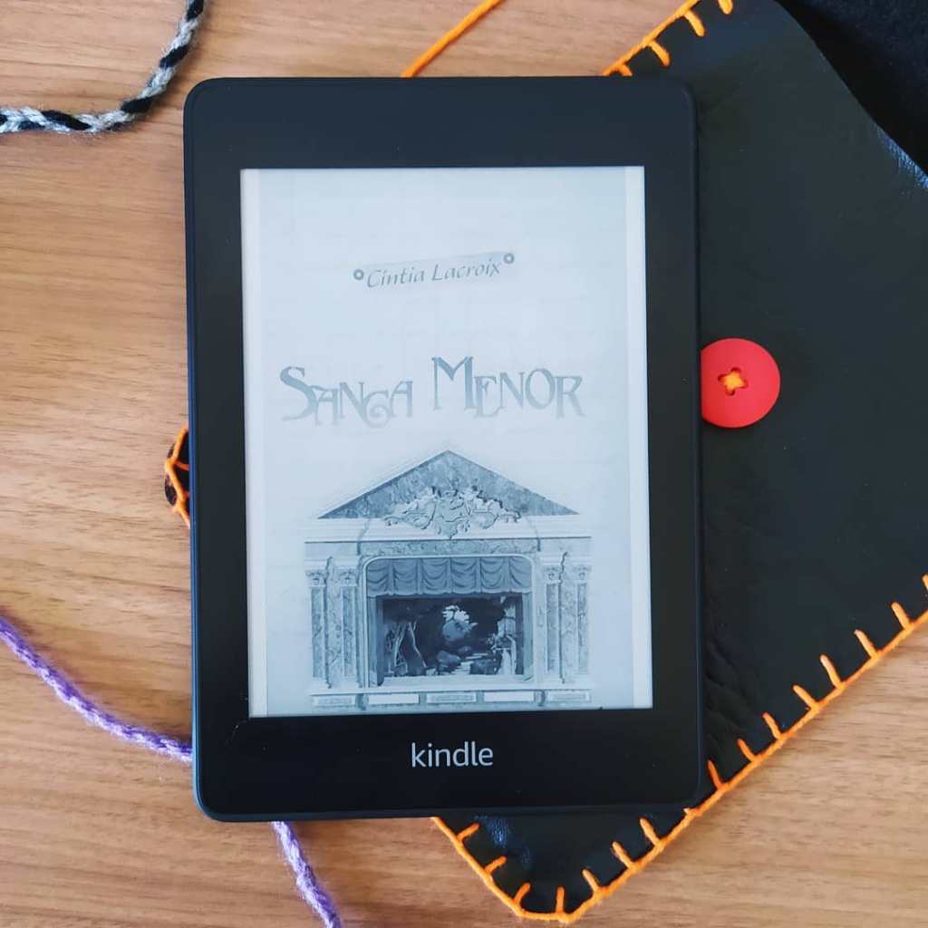 Kindle com a capa do livro Sanga Menor ao centro da imagem. O Kindle está posicionado acima de uma capa artesanal para kindle feita em courino preto, com botão vermelho e costura laranja. Há alguns fios de lã pela imagem, nas cores preta e cinza e roxa.