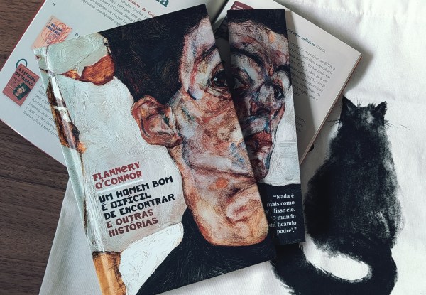 Capa do livro Um Homem Bom é Difícil de Encontrar, com o marcador de páginas personalizado do kit ao lado (à direita). O livro e o marcador estão em cima da revista co conteúdo complementar aberta, que por sua vez, está em cima da ecobag que veio como brinde no kit, mostrando o gato preto em sua estampa.