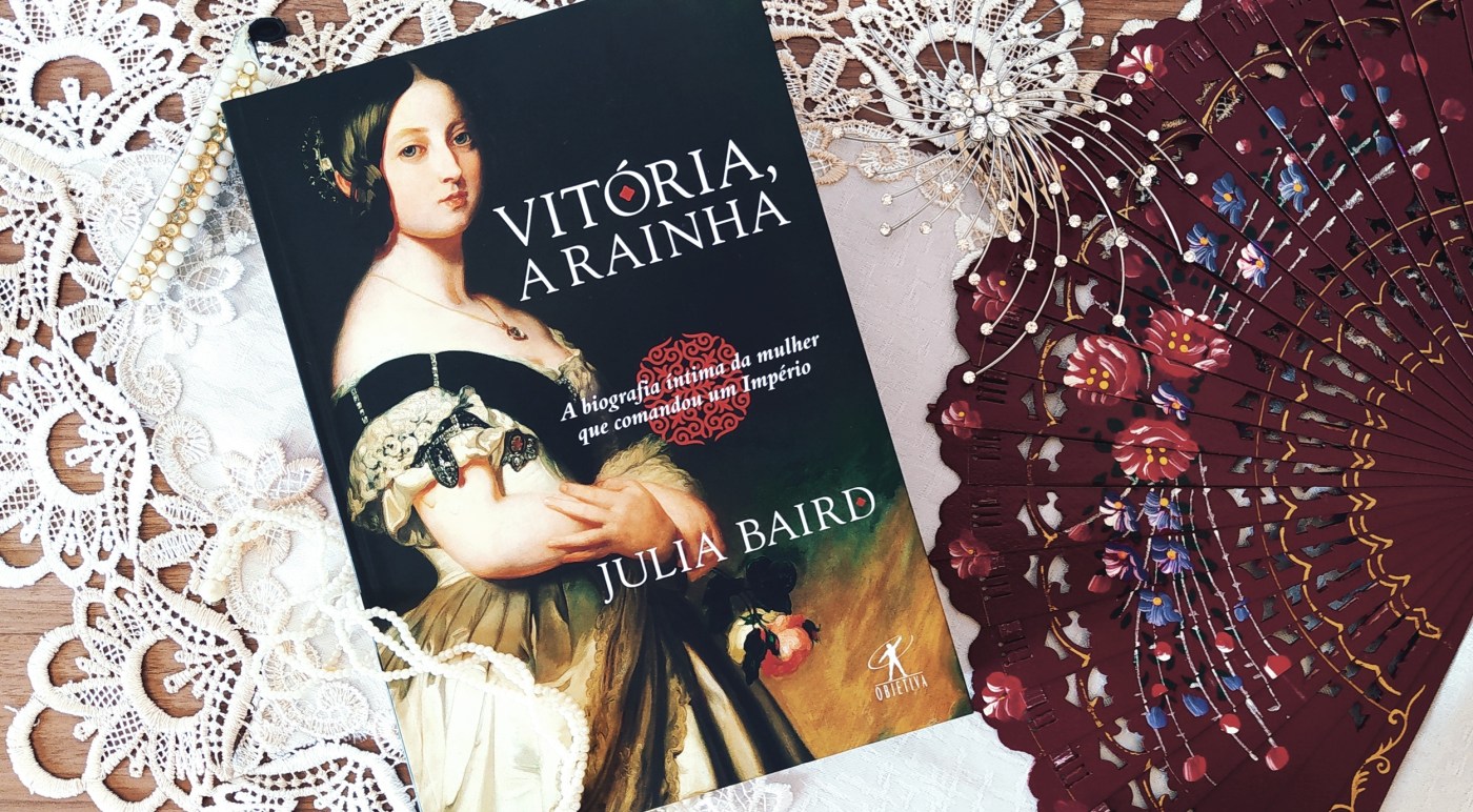 Livro Vitória, a Rainha, sobre uma toalha branca com bordas de renda. Há um leque envernizado com temas florais no lado direito da imagem, entre o leque e o livro. Na parte superior da imagem, há uma presilha de cabelo prateada com diversos strass. No lado esquerdo da imagem, no canto superior, há outra presilha de cabelo em formato circular, com fios saindo do centro e indo para as bordas, com imitações de pérolas e strass em formato linear. Há um colar de pérolas que passa parcialmente por cima do canto inferior esquerdo do livro.