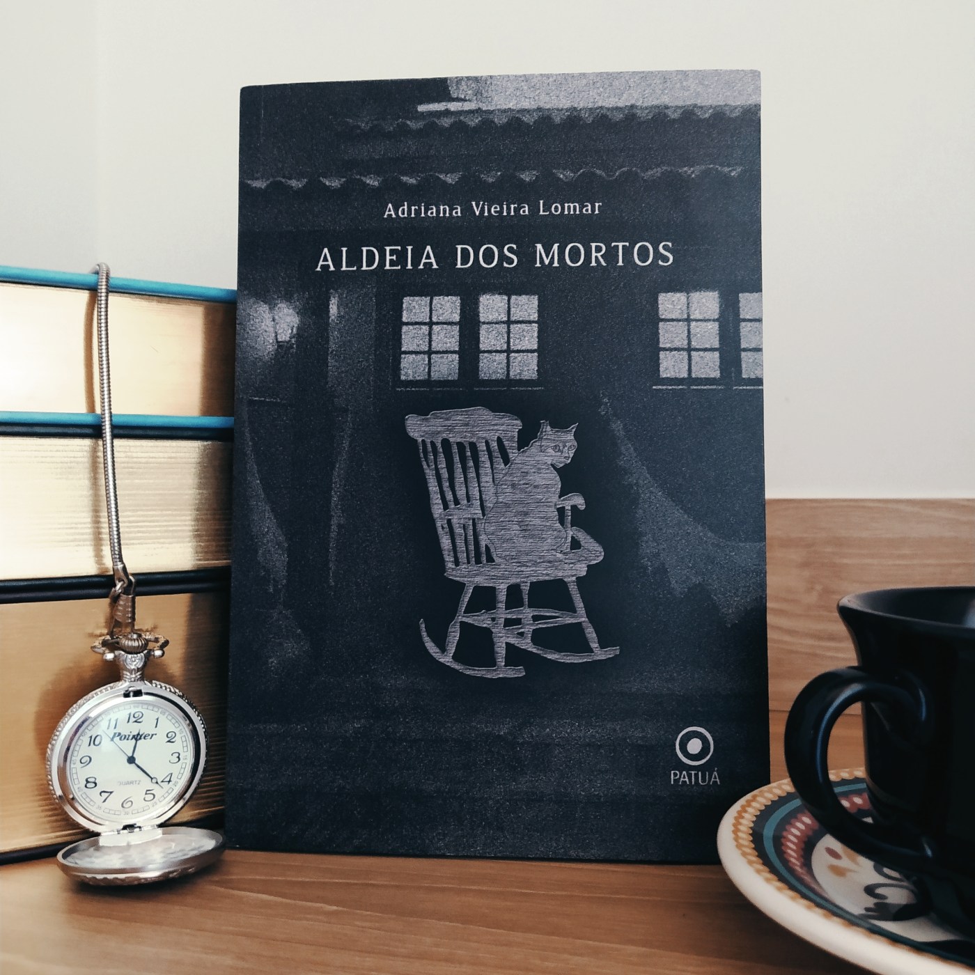 Livro Aldeia dos Mortos apoiado em uma pilha de livros, com um relógio de bolso aberto ao lado esquerdo e um pedaço de xícara em apoiada em um pires ao lado direito.