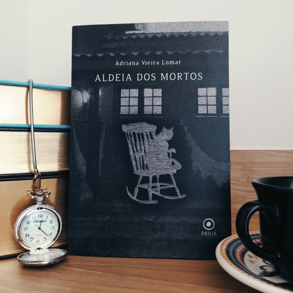 Livro Aldeia dos Mortos apoiado em uma pilha de livros, com um relógio de bolso aberto ao lado esquerdo e um pedaço de xícara em apoiada em um pires ao lado direito.