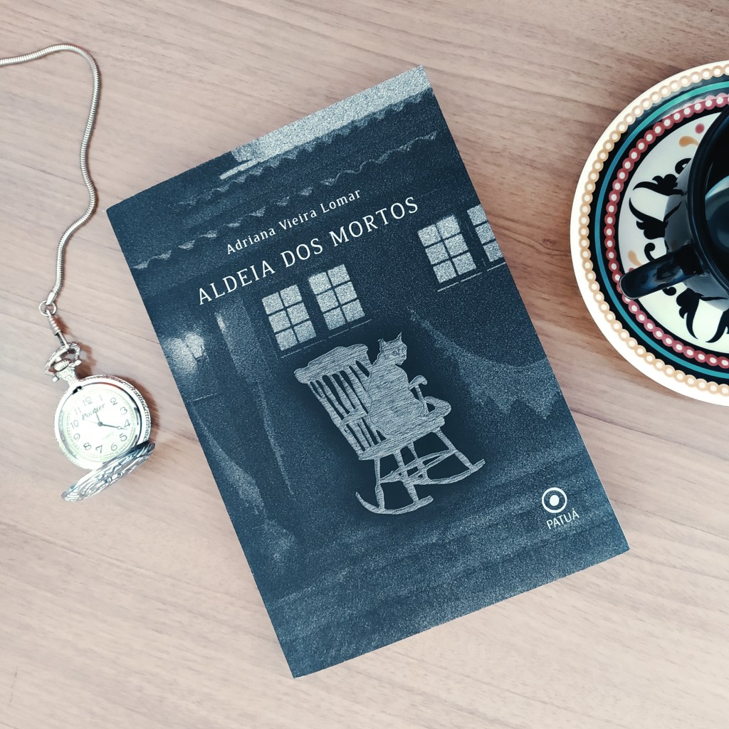 Livro Aldeia dos Mortos ao centro, com um relógio de bolso aberto à esquerda e uma parte de uma xícara acima de um pires à direita.