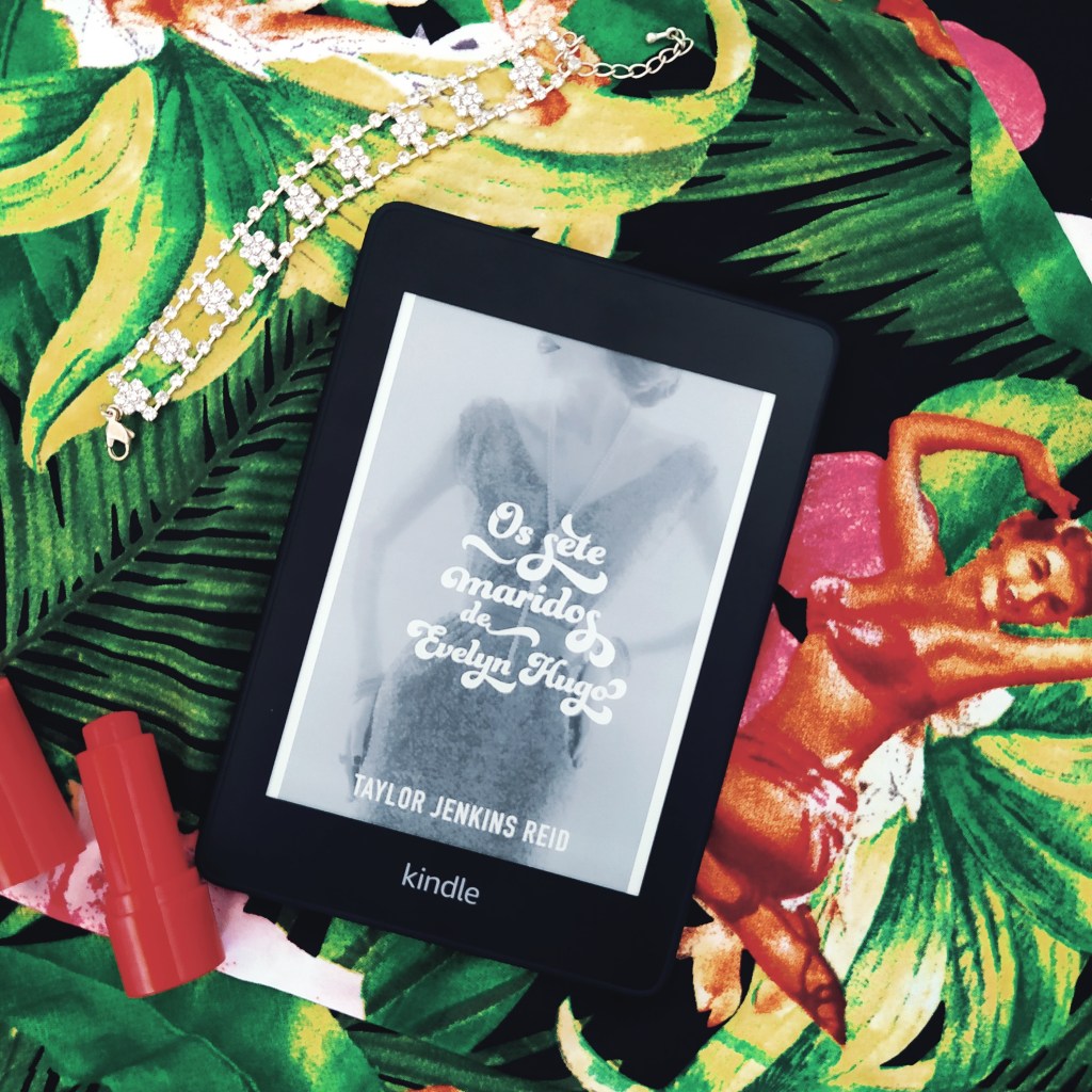 Kindle com a capa de Os Sete Maridos de Evelyn Hugo no centro da imagem. No canto superior esquerdo, há uma pulseira de zircônias. No canto inferior esquerdo, há um batom vermelho aberto. Ao fundo, há um tecido com tema floral e a imagem de uma pin-up.