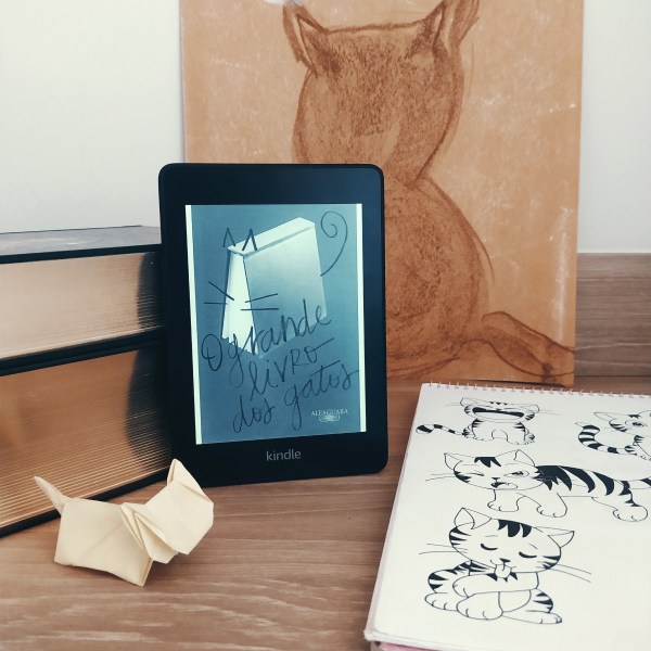 Kindle com a capa de O Grande Livro dos Gatos apoiado em uma pilha de livros, com uma pintura em giz à óleo em papel kraft da silhueta de um gato. À direita, há um caderno com desenhos em nanquim de gatos e à frente há um origami de gato.