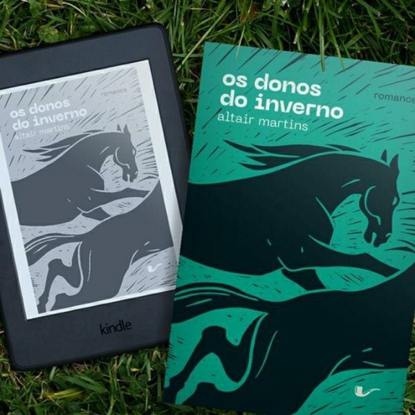 Kindle com a capa de Os Donos do Inverno à esquerda da imagem e a capa do exemplar físico do mesmo livro à direita. Ao fundo está um gramado.