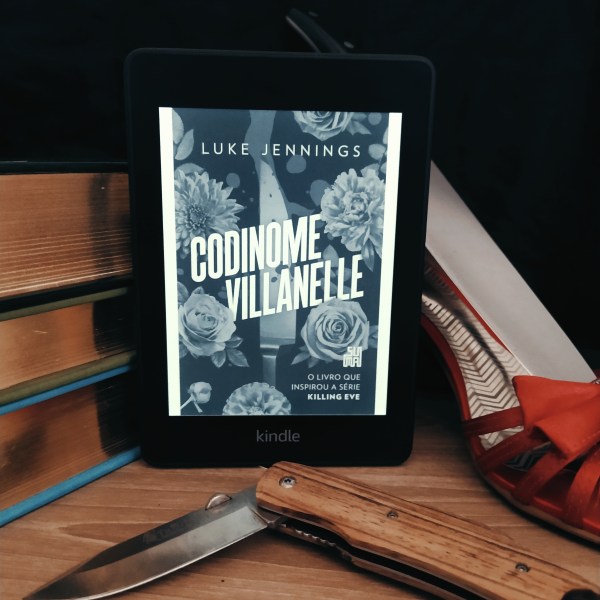 Kindle com a capa do livro Codinome Villanelle apoiado em uma pilha de livros. À direita, há uma sandália de salto alto vermelha com uma faca apoiada dentro dela. Abaixo, há um canivete aberto de forma a fazer um ângulo pouco maior que noventa graus.