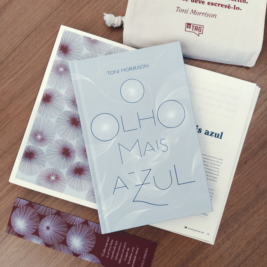 Exemplar de O Olho Mais Azul, posicionado em cima da revista aberta que vem no kit da TAG. Acima da composição está o brinde do kit, uma mini ecobag para livros, e abaixo está o marcador de páginas personalizado da TAG.