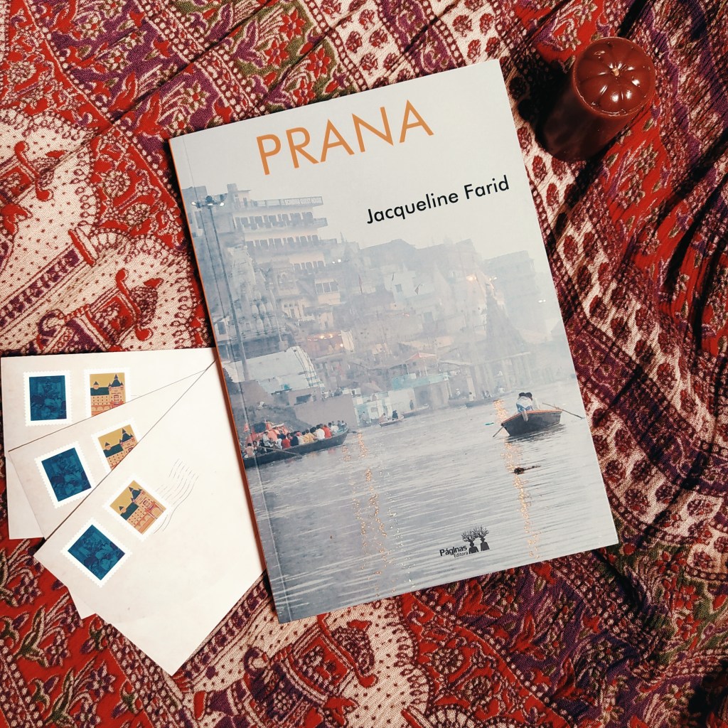Livro Prana, com uma vela à esquerda e acima e três envelopes de carta à direita e abaixo. Ao fundo, há um tecido com temas que remetem à Índia.