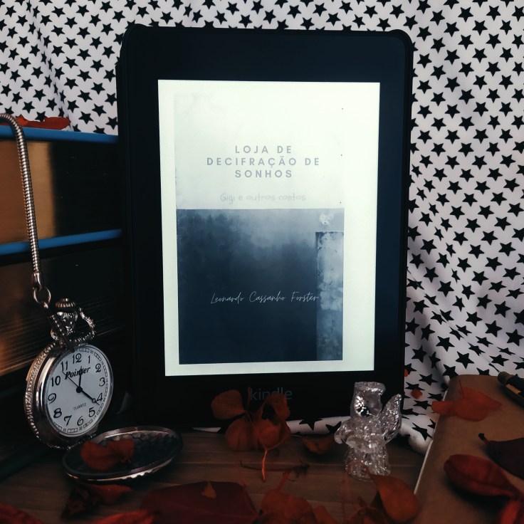 Kindle no centro da imagem com a capa do livro Loja de Decifração de Sonhos - Gigi e Outras Histórias. No lado esquerdo da imagem, há uma pilha de livros com um relógio de bolso aberto apoiado nela. No canto inferior esquerdo da imagem, há uma parte de uma caderneta com capa em kraft aparecendo. À frente do kindle, há um anjinho de cristal. Ao fundo, há um tecido branco estampado com estrelas pretas. Há folhas secas espalhadas pela superfície onde os objetos estão apoiados.