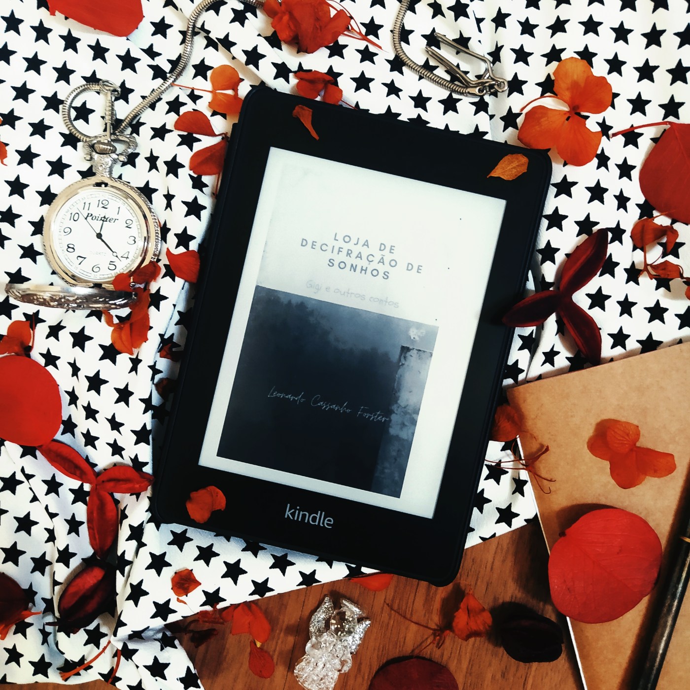 Kindle no centro da imagem com a capa do livro Loja de Decifração de Sonhos - Gigi e outras histórias. No canto inferior direito, há um pedaço de uma caderneta com capa em kraft e uma caneta em cima. No lado esquerdo, na metade superior, há um relógio de bolso prateado aberto, com o cordão passando por cima do kindle. No centro da parte inferior, há um anjinho de cristal. O fundo está quase totalmente coberto por um tecido branco com estampa de estrelas pretas. Há folhas secas espalhadas pela imagem.