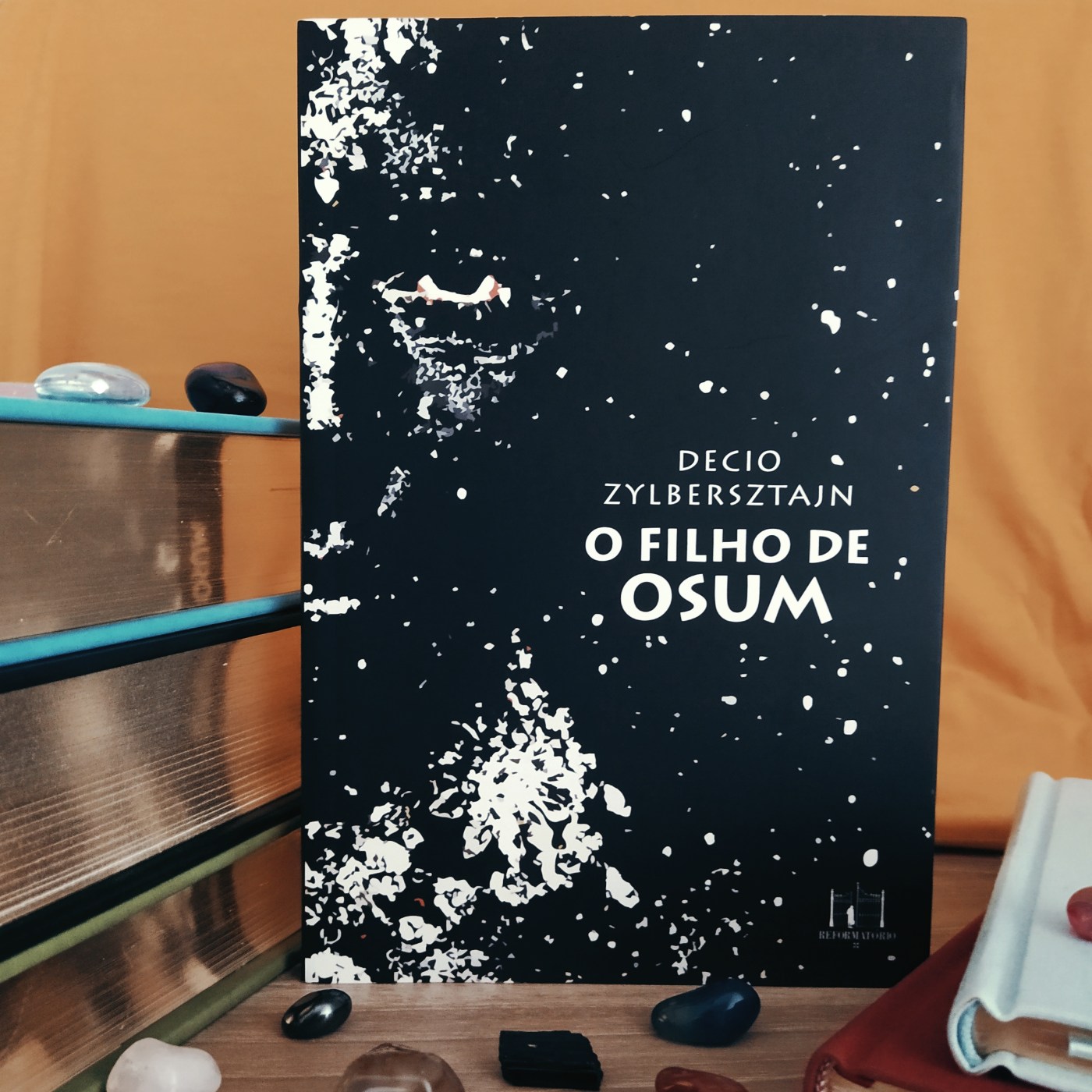 Livro O Filho de Osum em destaque no centro da imagem, com uma pilha de livros no lado esquerdo da imagem e dois livros pequenos com capa em tecido no lado direito da imagem. o fundo é amarelo e há pedras ornamentais espalhadas pelas superfícies dos livros e onde estes estão apoiados.