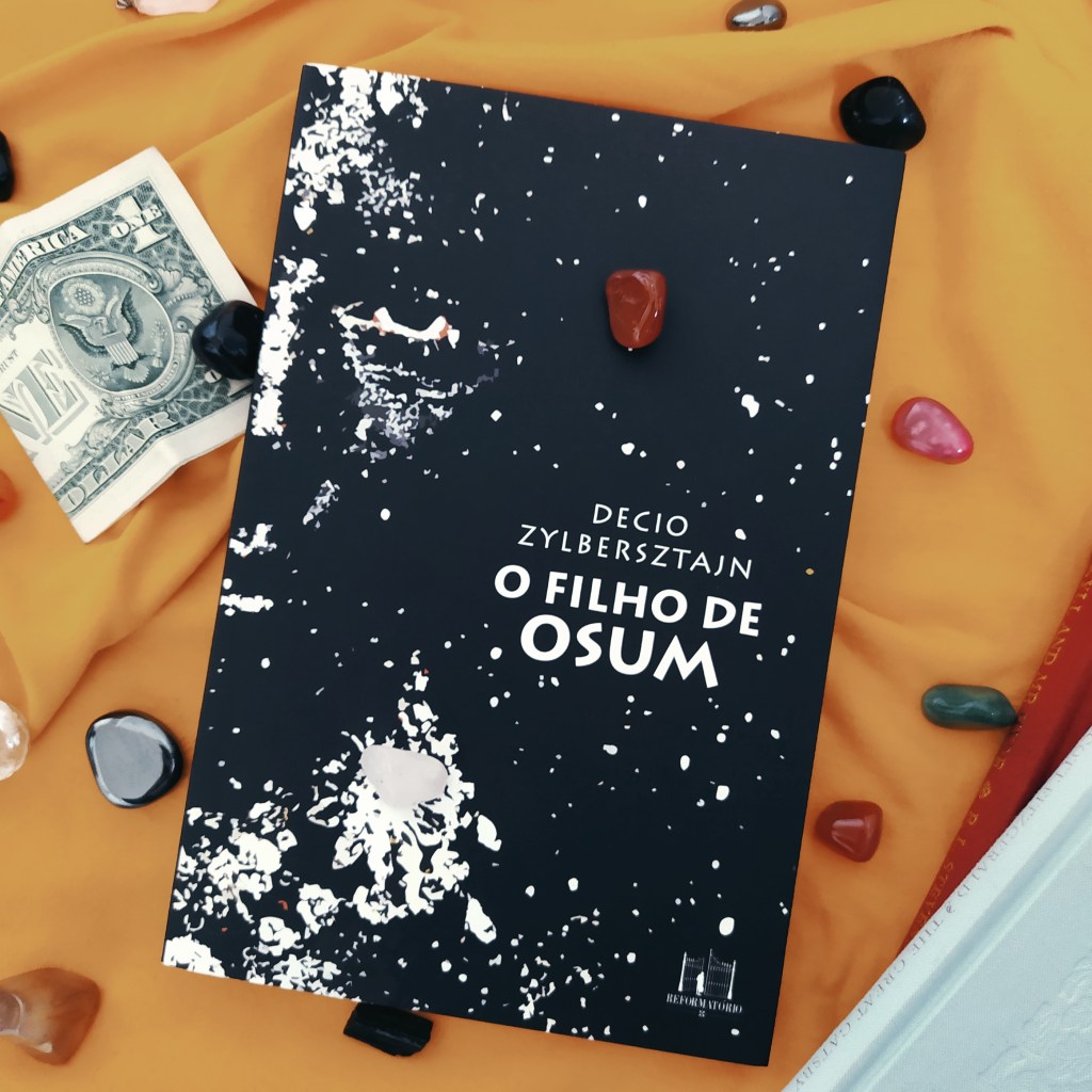 Livro O Filho de Osum no centro da imagem, com dois livros com capa em tecido no canto inferior direito da imagem, uma nota de um dólar na parte superior esquerda da imagem e pedras ornamentais pela imagem. O fundo é amarelo.
