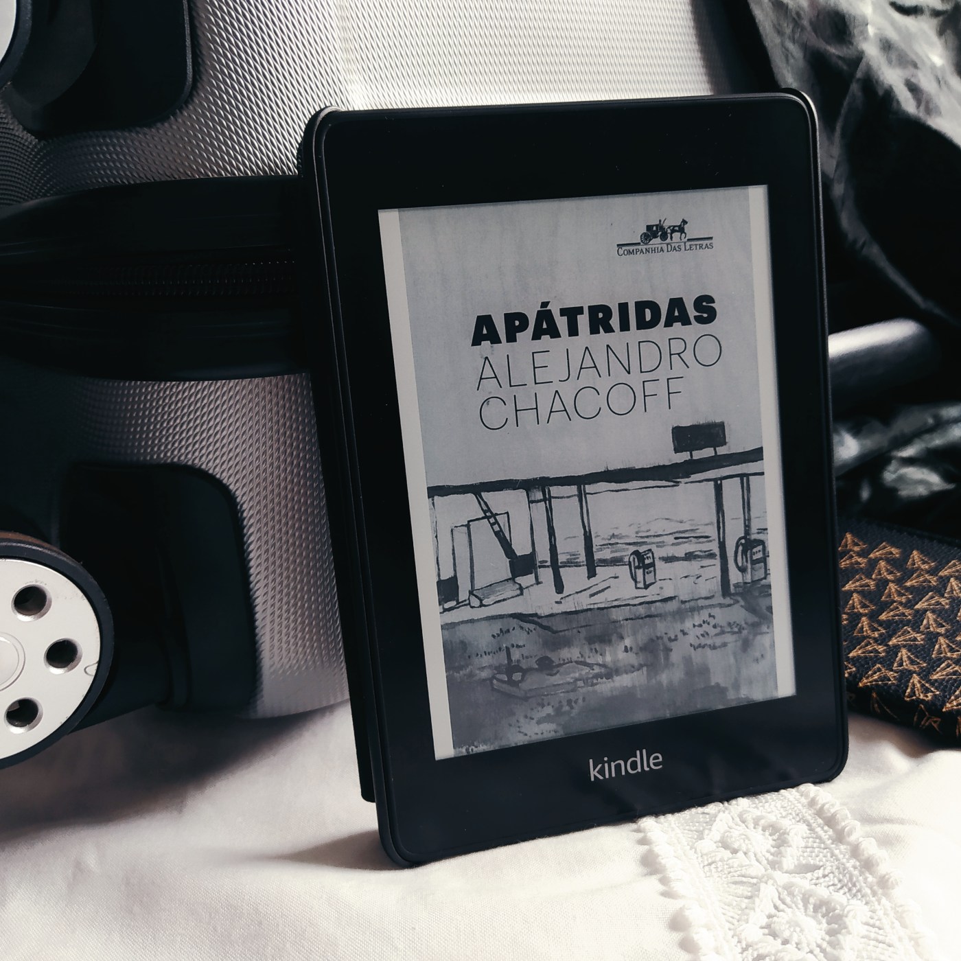 Kindle no centro da imagem com a capa do livro Apátridas, apoiado em uma mala de rodinhas cinza com detalhes em preto, sobre um tecido branco. Ao lado, um livreto com capa de passaporte, com estampa de aviões de papel.