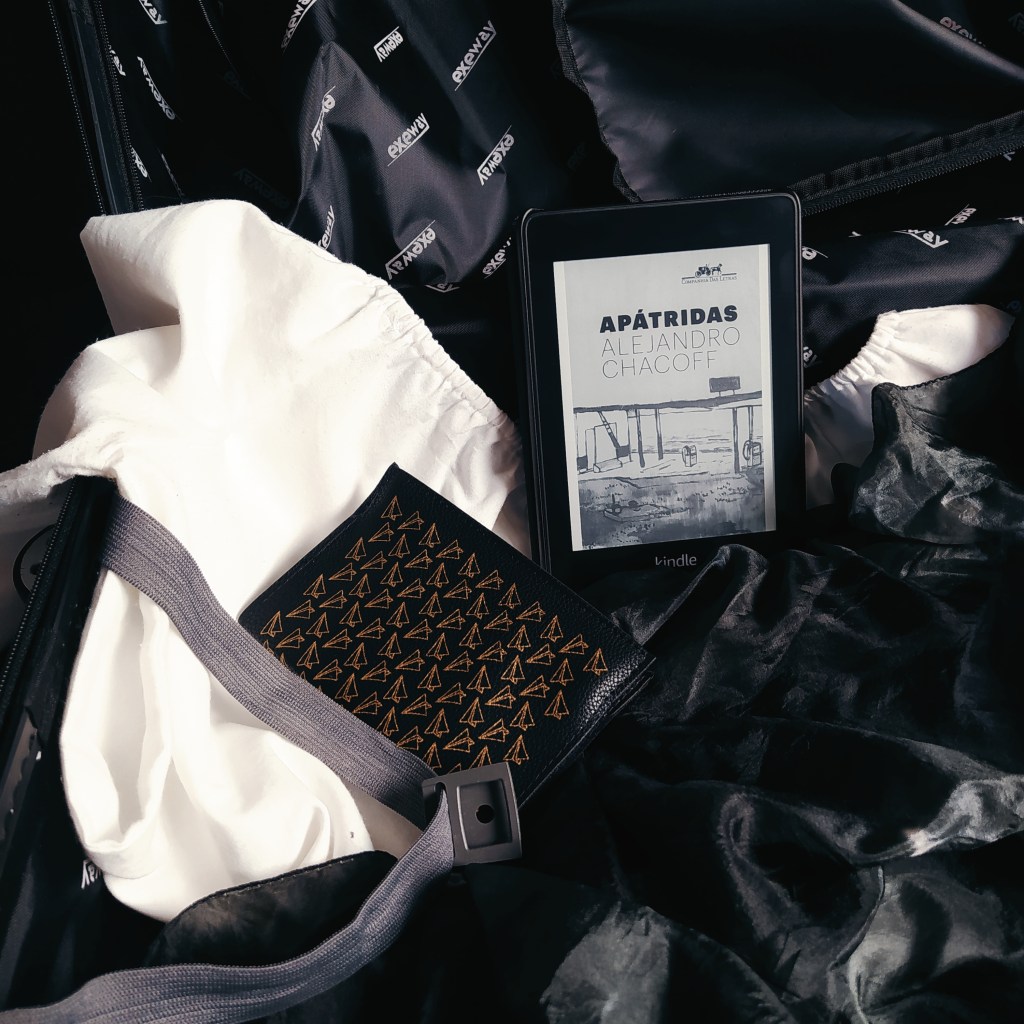 Mala de viagem aberta, com uma camiseta de manga comprida branca aberta com a manga para fora, um tecido preto jogado dentro da mala, um livreto com capa de passaporte com estampa de aviões de papel e um kindle com a capa do livro Apátridas.