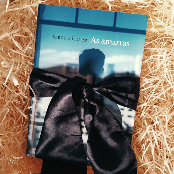 Livro As Amarras no centro da imagem, com uma faixa preta em torno dele fazendo um laço. O fundo é de palha.