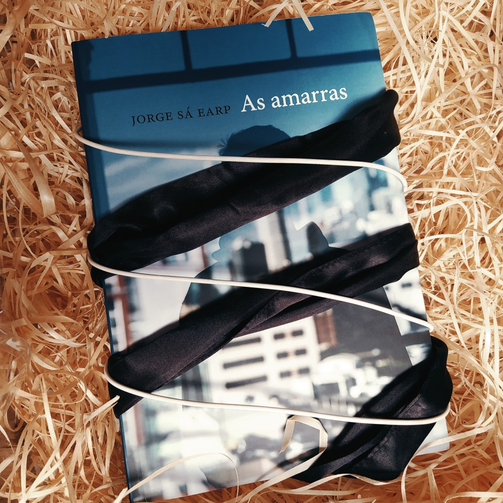 Livro As Amarras no centro da imagem, com uma faixa preta e um fio branco enrolados nele como se estivesse amarrado. O fundo é de palha.