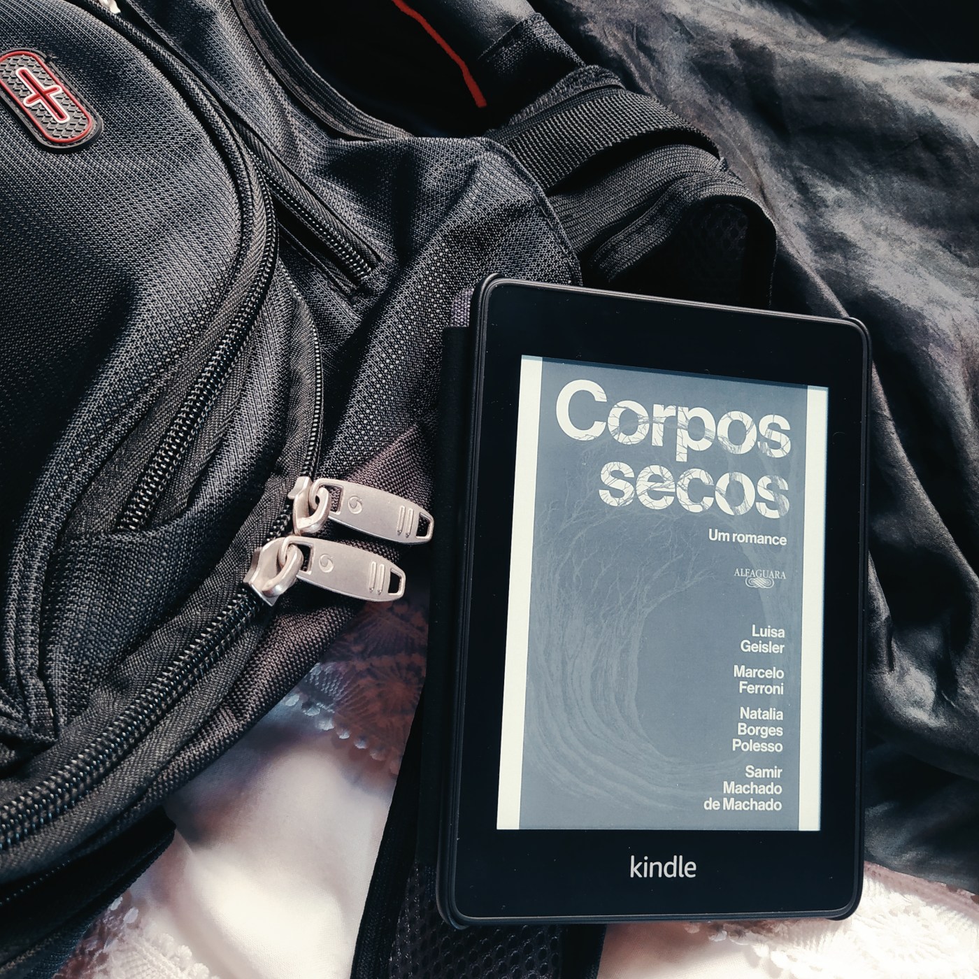 Kindle com a capa do livro Corpos Secos, apoiado em uma mochila preta, que está na metade esquerda da imagem. O fundo é composto de tecidos preto, na parte superior, e branco, na parte inferior.