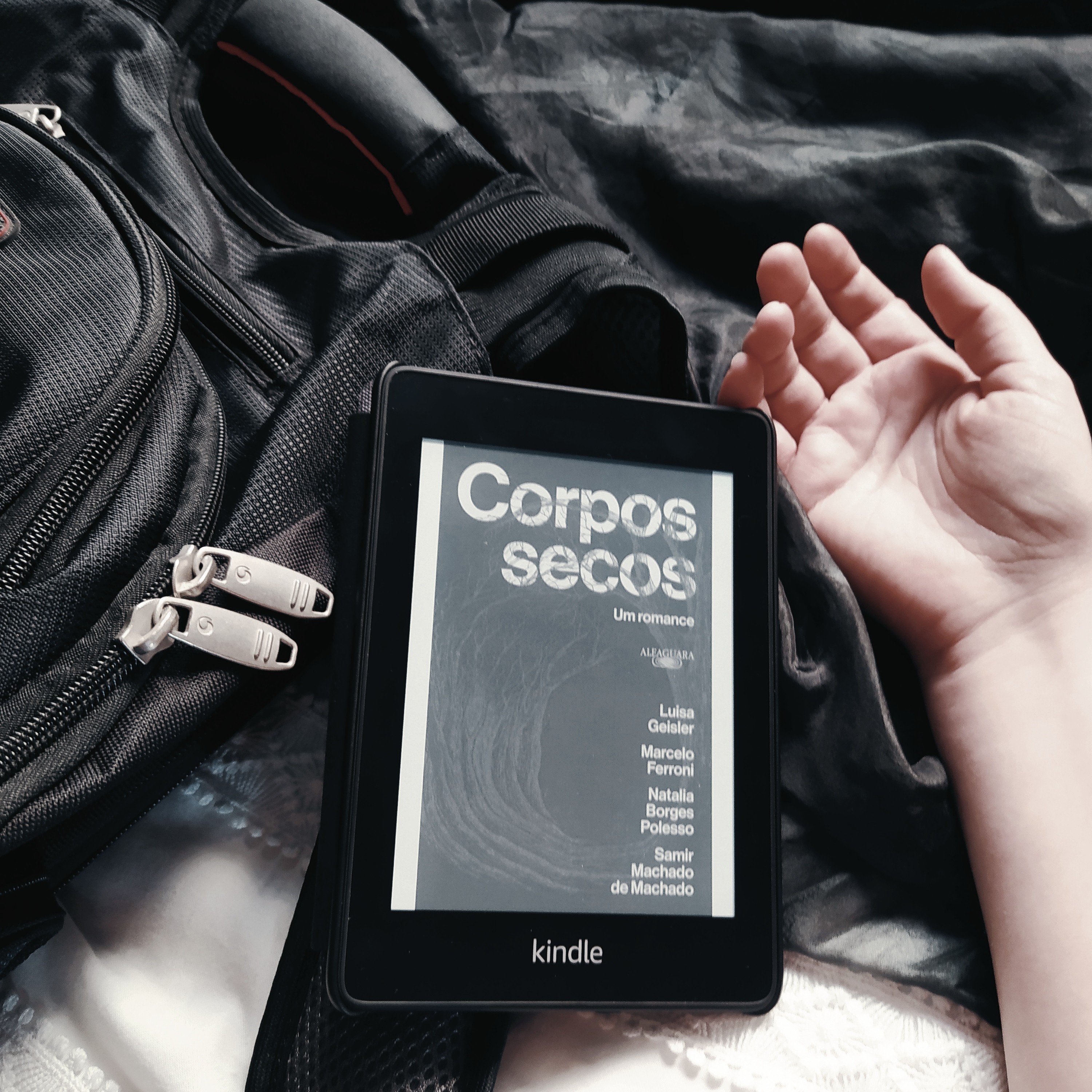 Kindle com a capa do livro Corpos Secos, apoiado em uma mochila preta, que está na lateral esquerda da imagem. Há uma mão caída ao lado do kindle, na parte direita da imagem. O fundo é composto de tecidos preto, na parte superior, e branco, na parte inferior.