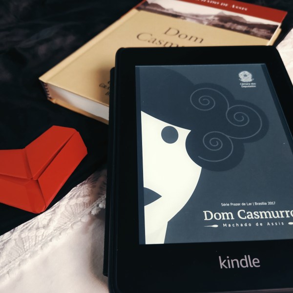 Kindle em primeiro plano com a capa do livro Dom Casmurro. Ao fundo, há um livro físico de Dom Casmurro, onde o kindle se apoia. No lado esquerdo da foto, mais ao centro, há um origami de coração vermelho. O fundo é feito com tecidos preto e branco.