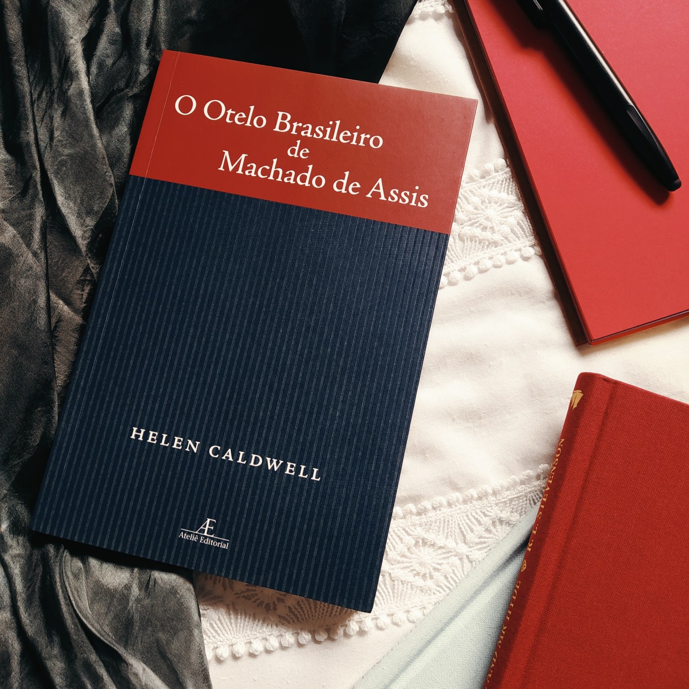 Livro O Otelo Brasileiro de Machado de Assis na parte centralizada mais à esquerda da imagem, com um bloco de anotações de folhas vermelhas no canto superior direito, com uma caneta preta por cima. No canto inferior esquerdo, há dois livros de capa de tecido, um por cima do outro, sendo azul o que está embaixo e vermelho o que está em cima. O fundo é branco, com um tecido preto na lateral esquerda.
