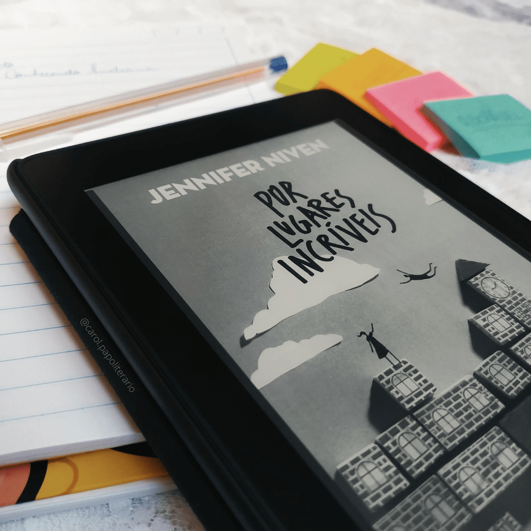 Kindle em primeiro plano, com a capa do livro Por Lugares Incríveis. O Kindle está apoiado em um caderno pautado. Há uma caneta bic em cima do caderno, mais ao fundo. Também é possível ver, ao fundo e desfocado, ao lado do caderno, 4 bloquinhos de notas autoadesivas.