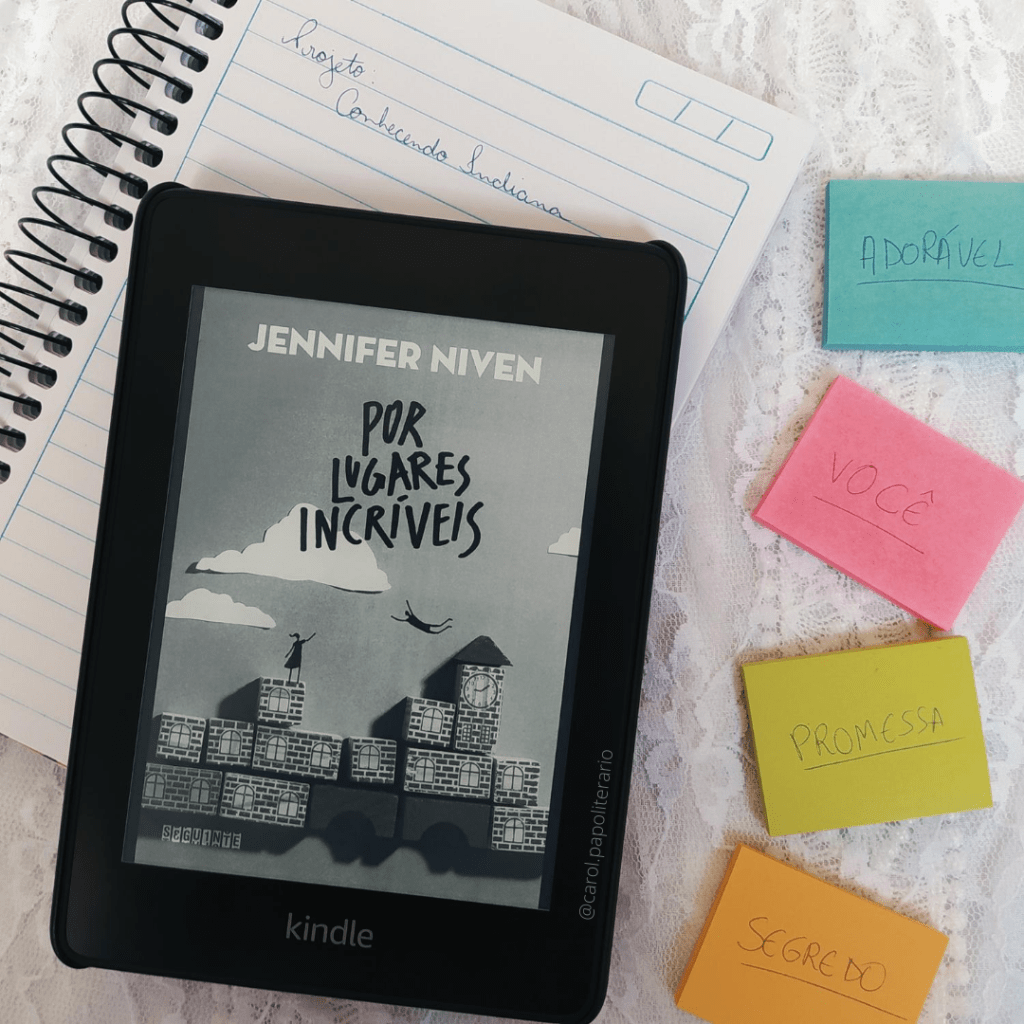 Kindle com a capa do livro Por Lugares Incríveis ocupando a maior parte do lado esquerdo da foto. O Kindle está apoiado em um caderno com espiral e pautado, onde se lê: Projeto Conhecendo Indiana. Ao lado da composição, estão 4 bloquinhos de notas autoadesivas, onde se lê, de cima para baixo: adorável, você, promessa e segredo.