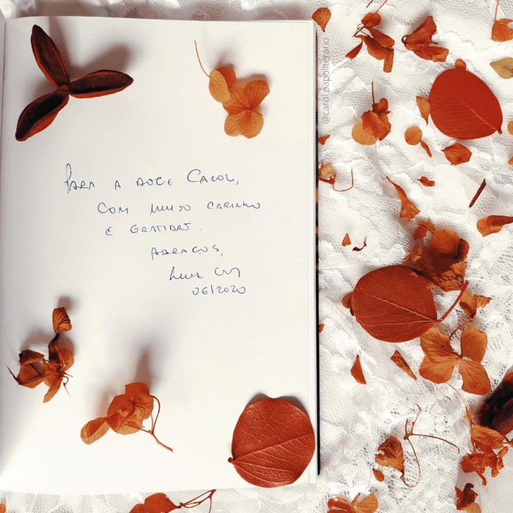 No terço esquerdo da imagem, há um livro aberto em uma dedicatória, onde se lê: "Para a doce Carol, com muito carinho e gratidão. Abraços, Luiz Eudes. 06/2020". Há folhas secas por toda a imagem e o fundo é composto por um tecido rendado branco.