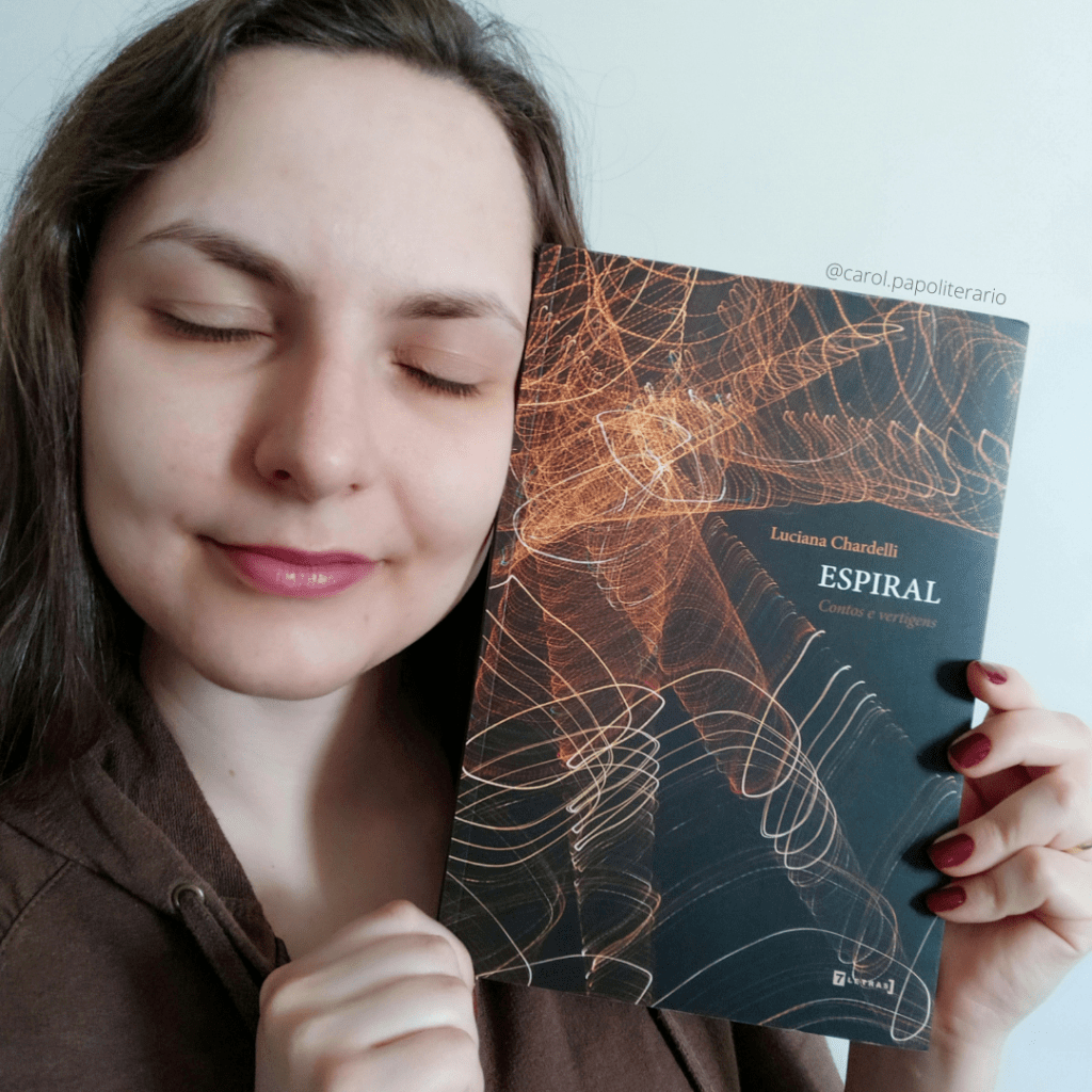 Carol segurando o livro Espiral próximo ao rosto. Ela está com os olhos fechados e um leve sorriso.
