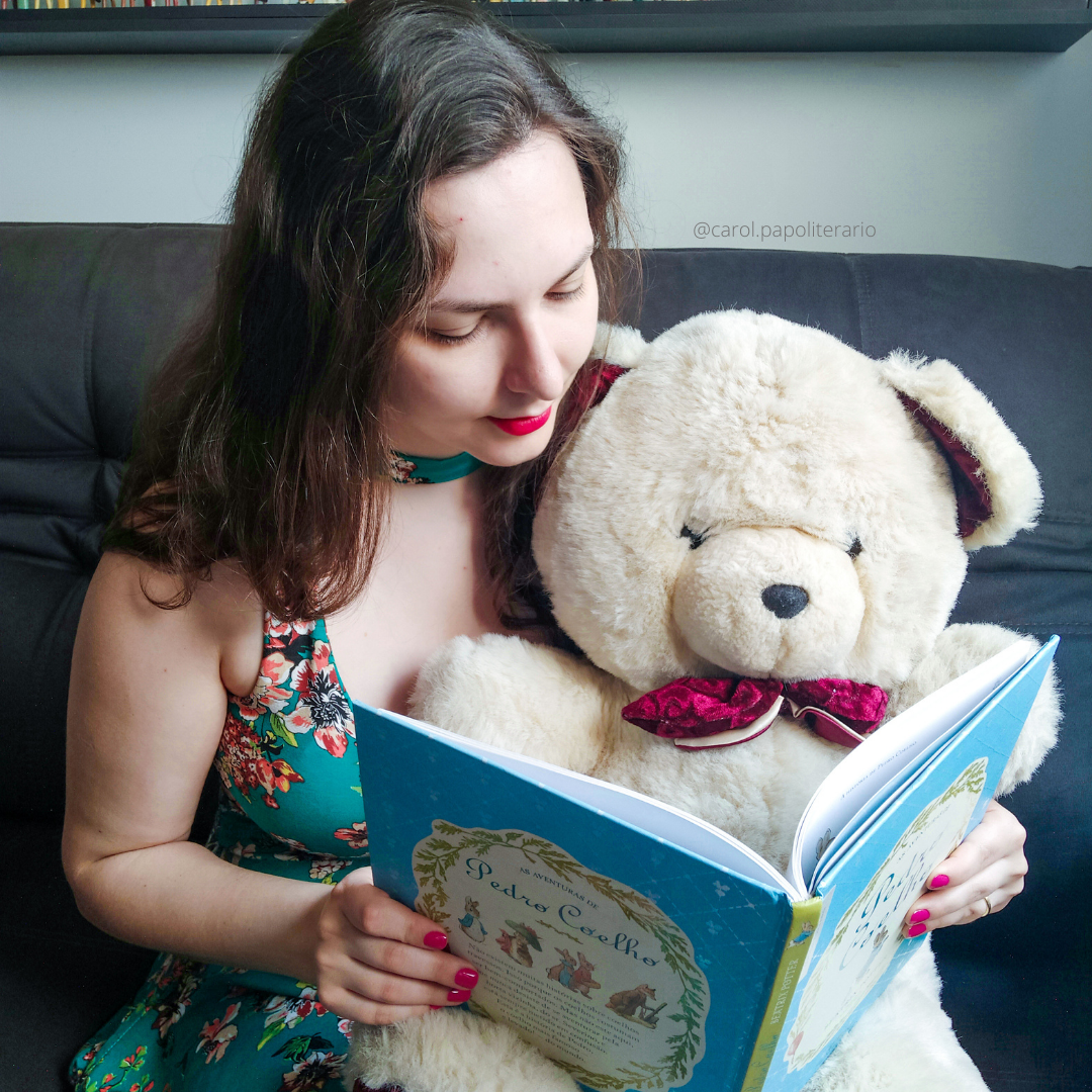 Moça sentada no sofá com um urso de pelúcia no colo. Ela está com um livro infantil aberto, como se estivesse lendo para o urso.