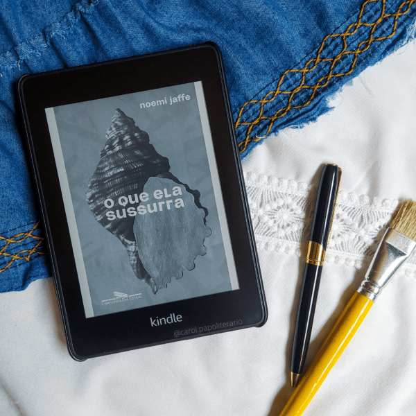 Kindle com a capa do livro O Que Ela Sussura, com uma caneta e um pincel no canto inferior direito da imagem.