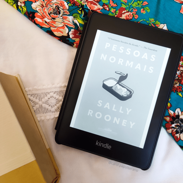 Kindle com a capa do livro de Pessoas Normais no centro da imagem, levemente deslocado para a direita. No canto superior direito, há um tecido com tema florido, onde o kindle se apoia parcialmente. No canto inferior esquerdo, há um livro de capa lisa aparecendo parcialmente.