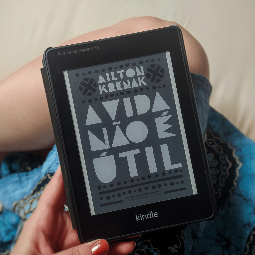 Carol segurando o Kindle com a capa do livro A Vida não é Útil. Aparece na foto um pedaço da perna e a barra de um vestido.