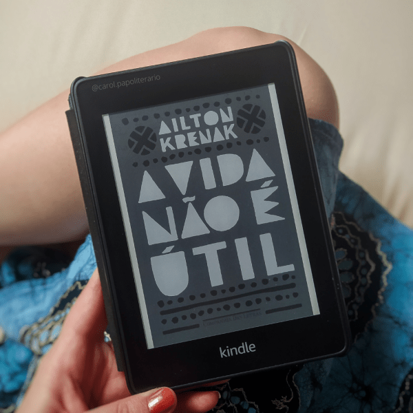 Carol segurando o Kindle com a capa do livro A Vida não é Útil. Aparece na foto um pedaço da perna e a barra de um vestido.