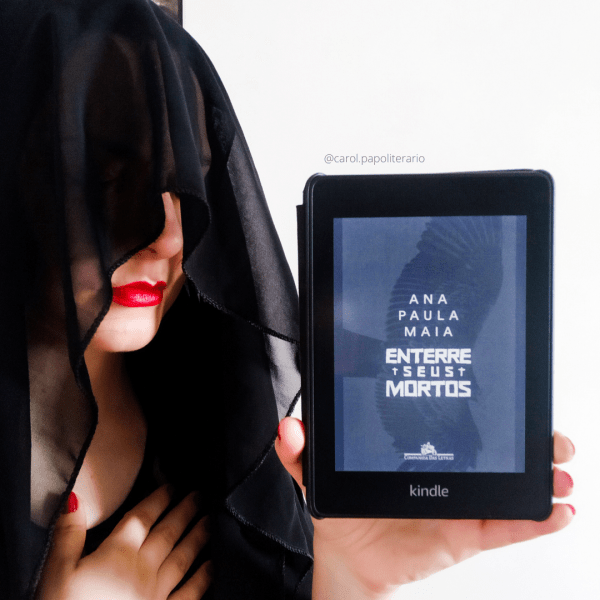 Carol com um véu preto cobrindo o rosto, deixando apenas parte do nariz e a boca à mostra. Ela segura um Kindle com a capa do livro Enterre Seus Mortos.