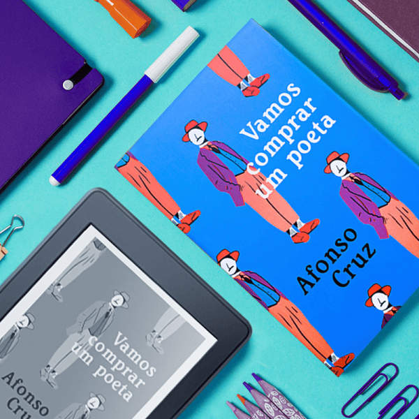 Livro Vamos Comprar um Poeta, ao lado de um Kindle (em 90 graus) com a capa do mesmo livro. Ao redor existem vários objetos de papelaria, como caneta, clips e lápis de cor.