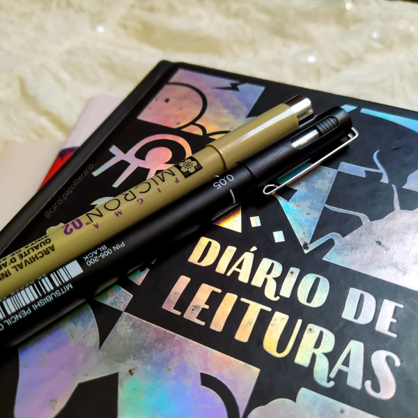 Diário de leituras com duas canetas em cima da capa e um livro por baixo, aparecendo apenas um pedaço da lombada, não sendo possível identificar qual é o livro.