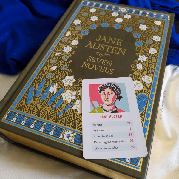 Livro de Jane Asuten, edição capa dura em inglês, contendo os sete romances da autora. Em cima do livro, há uma carta de jogo com um desenho representando Jane Austen, apresentando algumas habilidades logo abaixo, sendo elas: Vendas: 77. Prêmios: 15. Impacto Social: 86. Personagens marcantes: 92. Livros publicados: 30.