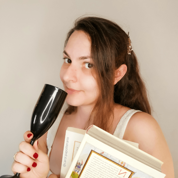 Carol com uma mecha do cabelo preso na lateral, segurando dois livros em uma mão e uma taça na outra, erguida na altura da boca.