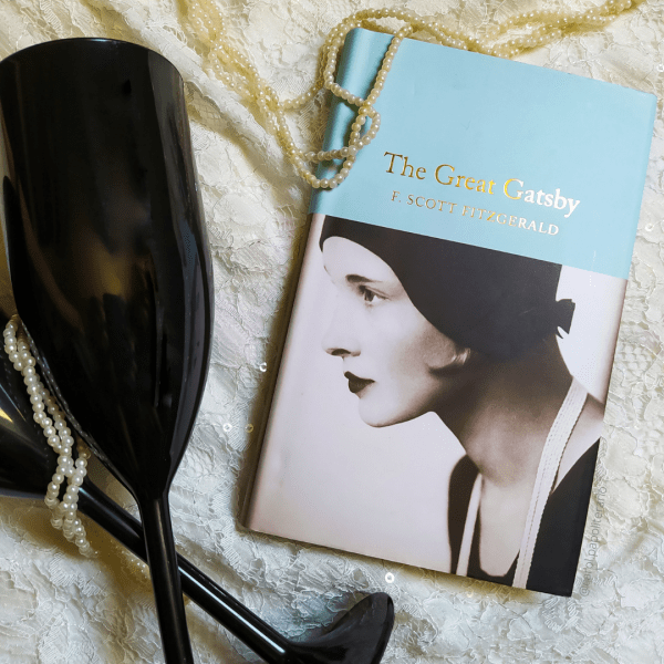 Livro O Grande Gatsby em edição em inglês, com um colar de pérolas passando pela parte superior do exemplar. Há duas taças cruzadas ao lado do livro, à esquerda.