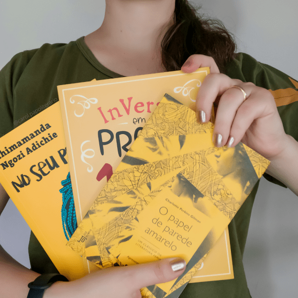 Carol segurando três livros de contos à sua frente, em forma de leque, sendo eles: No seu pescoço, InVerso em Prosa: 16 anos em muitas histórias, e O papel de parede amarelo.
