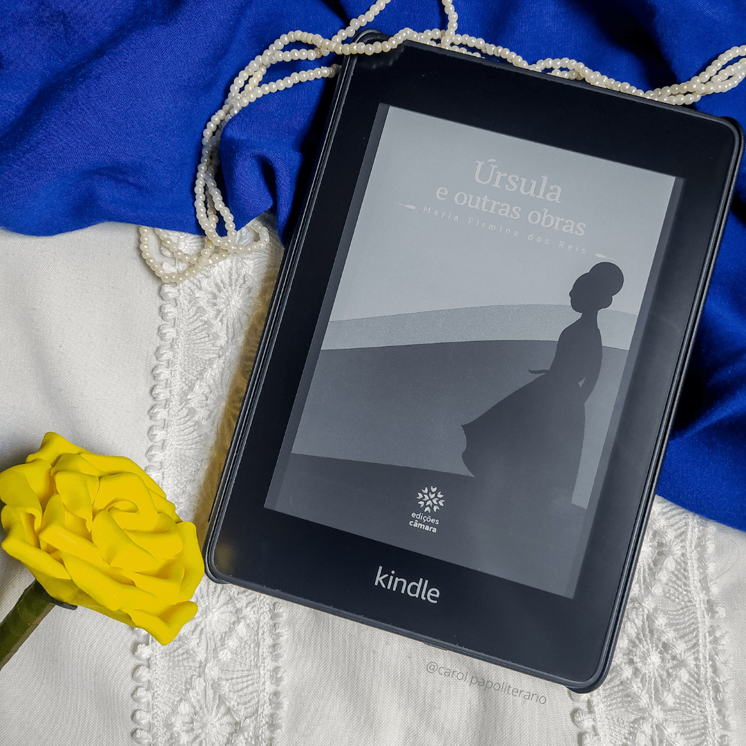 Kindle com a capa do livro Úrsula, com um colar imitando pérolar passando pela parte superior do aparelho e uma rosa artificial amarela ao lado do aparelho, no canto inferior esquerdo da imagem.
