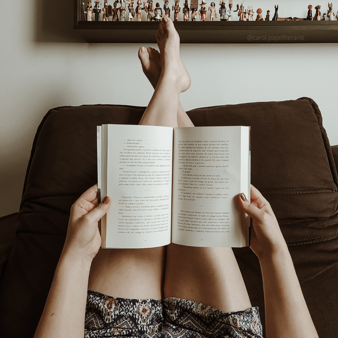 Carol deitada no sofá, com as pernas para cima, apoiadas no encosto, com um livro aberto. Na imagem, aparece apenas as pernas e as mãos segurando o livro.