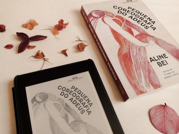 Livro "Pequena coreografia do adeus" ao lado de um kindle com a capa do mesmo título ao lado, dispostos em diagonal (canto inferior esquerdo ao canto superior direito), com folhas secas em torno dos objetos.