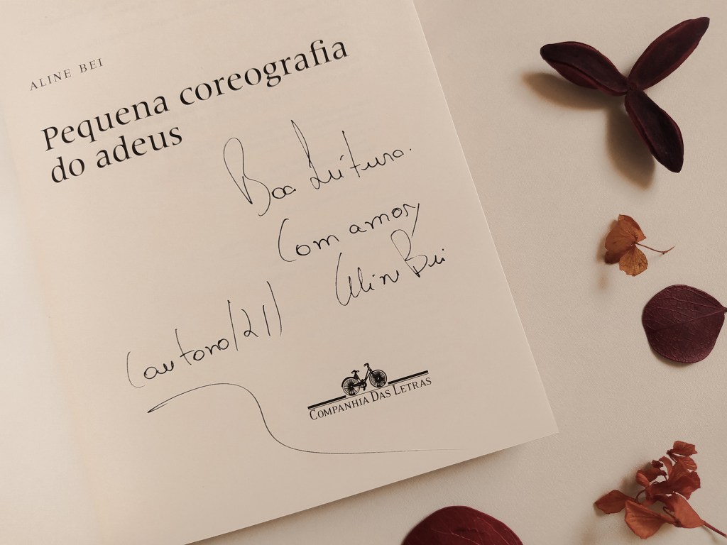 Livro "Pequena coreografia do adeus" aberta na folha de rosto, com o autógrafo da autora, onde se lê: "Boa leitura. Com amor, Aline Bei (outono/21)". Há folhas secas ao redor do livro.