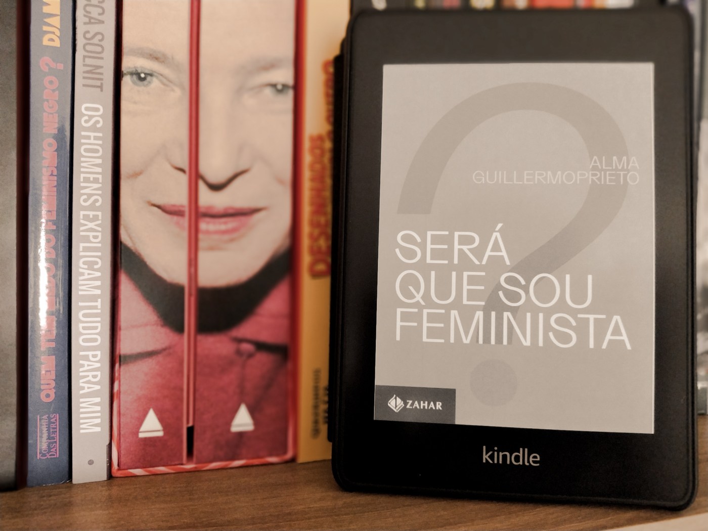 Kindle com a capa do livro "será que sou feminista?" apoiado em pé em cima de uma prateleira de livros. Ao fundo, há livros de autoras feministas, como Simone de Beauvoir e Djamila Ribeiro.