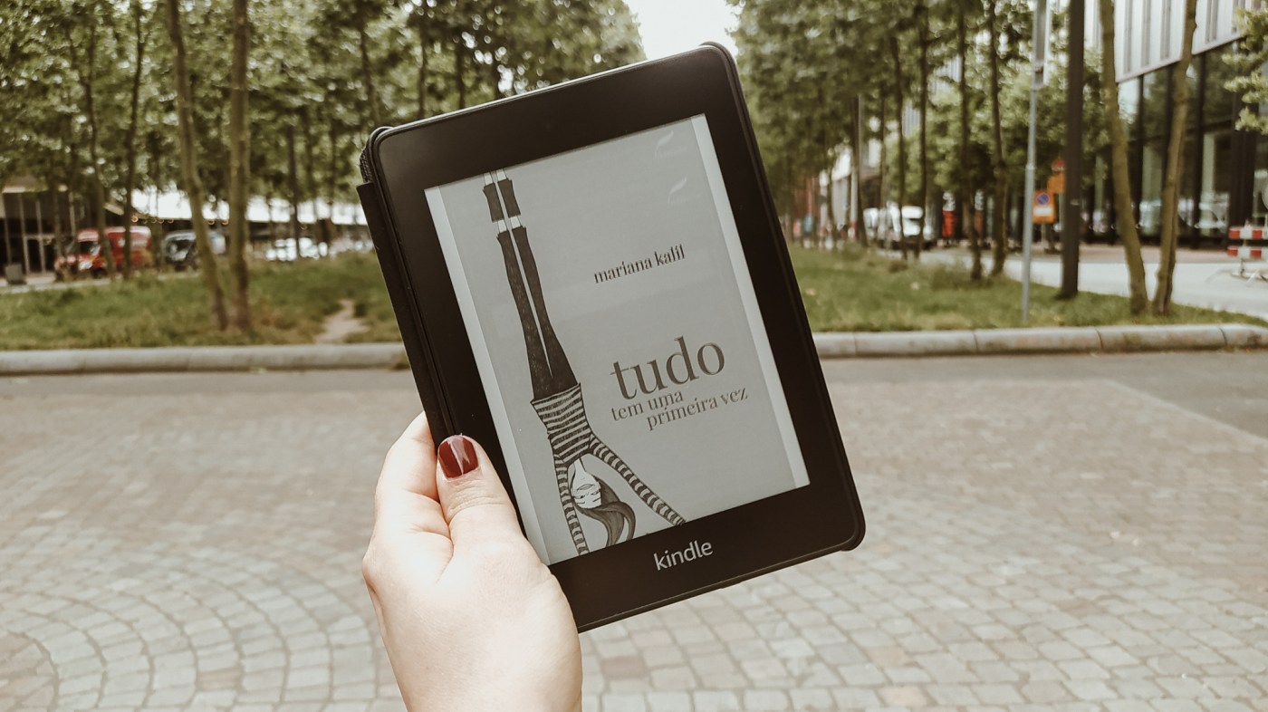 Mão da Carol segurando um Kindle com a capa do livro "Tudo tem uma primeira vez". Ao fundo, desfocado, estão árvores de um parque.